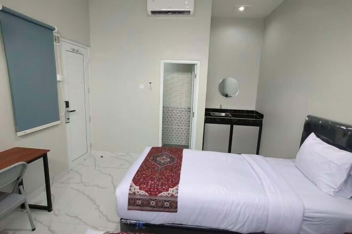 A hotel room with a bed, a desk, a chair, a mirror, a door, and an air conditioning unit.