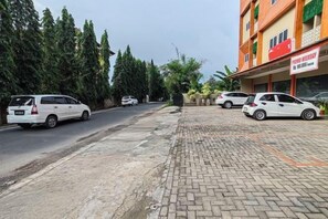 A street view with parked cars, a building with a sign, and a paved area. - Joy Hotel (Rajabasa)
