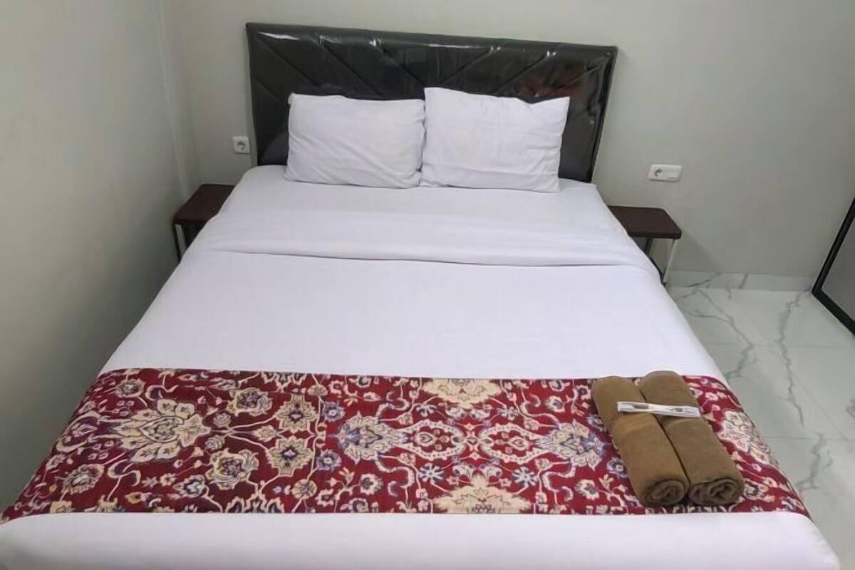 A bed with a headboard, two pillows, a red patterned bedspread, and a rolled towel on the footboard.