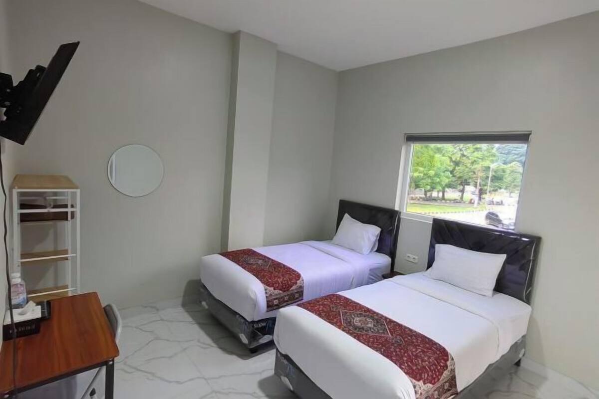 A hotel room with two beds, a desk, a chair, a TV, a mirror, and a window with a view of trees.
