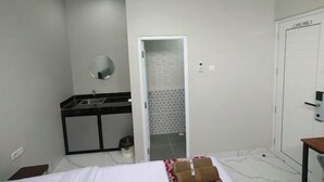 Habitación de hotel con cama, mesita de noche pequeña, espejo, baño con lavamanos y ducha, y una puerta.
