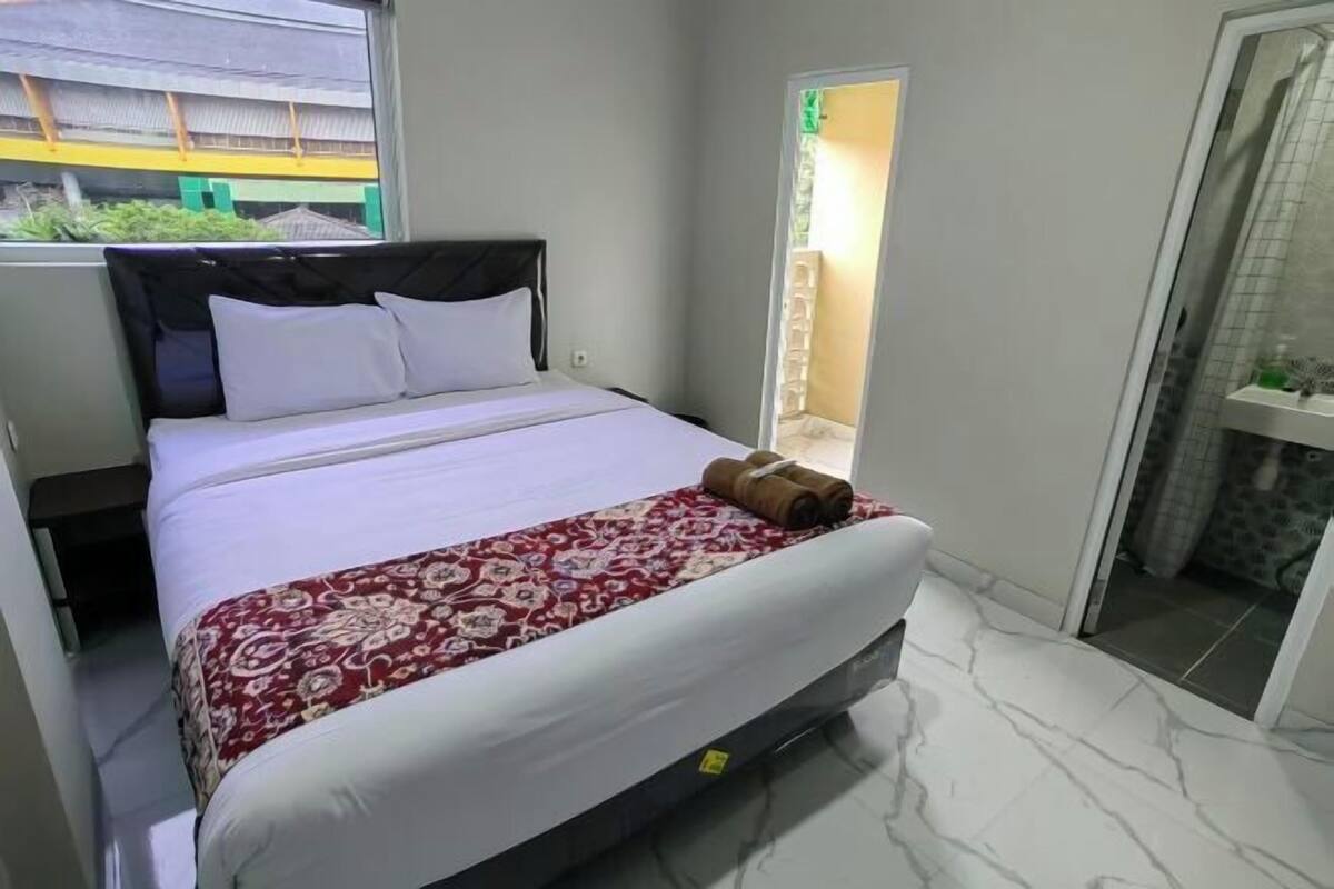A hotel room with a bed, a window with a view of a building, a door leading to a bathroom, and a marble floor.