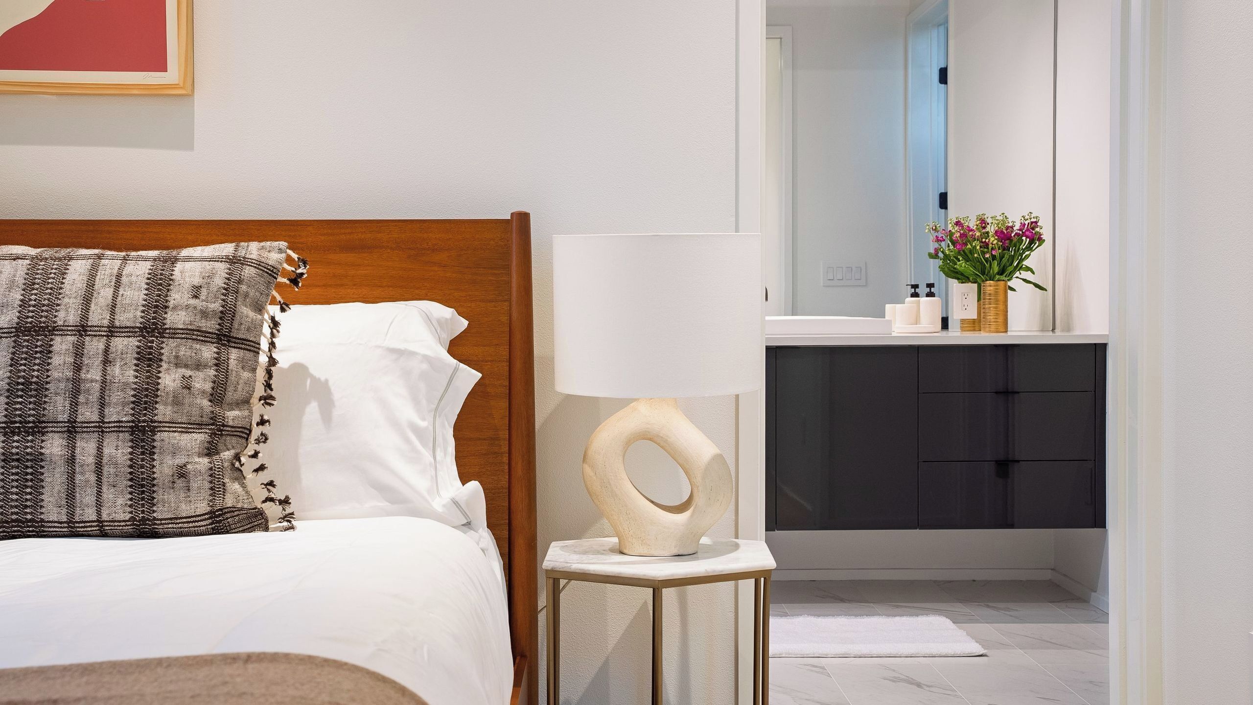 Ein ordentlich bezogenes Bett mit einem gemusterten Kopfkissen, einer Nachttischlampe und Blick in ein Badezimmer mit Waschbecken und Pflegeprodukten.