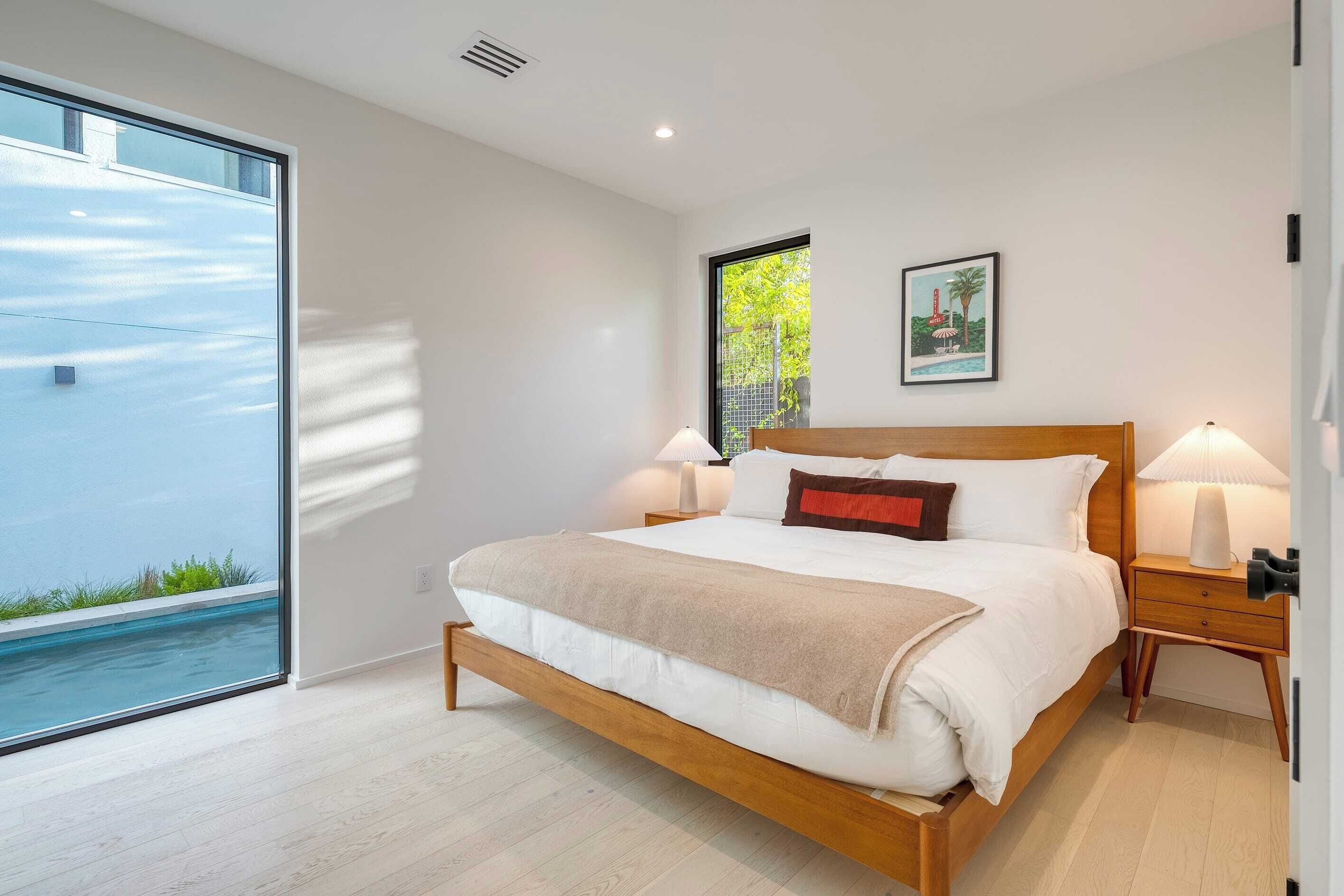 Un dormitorio moderno con una cama grande, lámparas de noche, una mesita de noche de madera y vistas a una piscina a través de un ventanal.