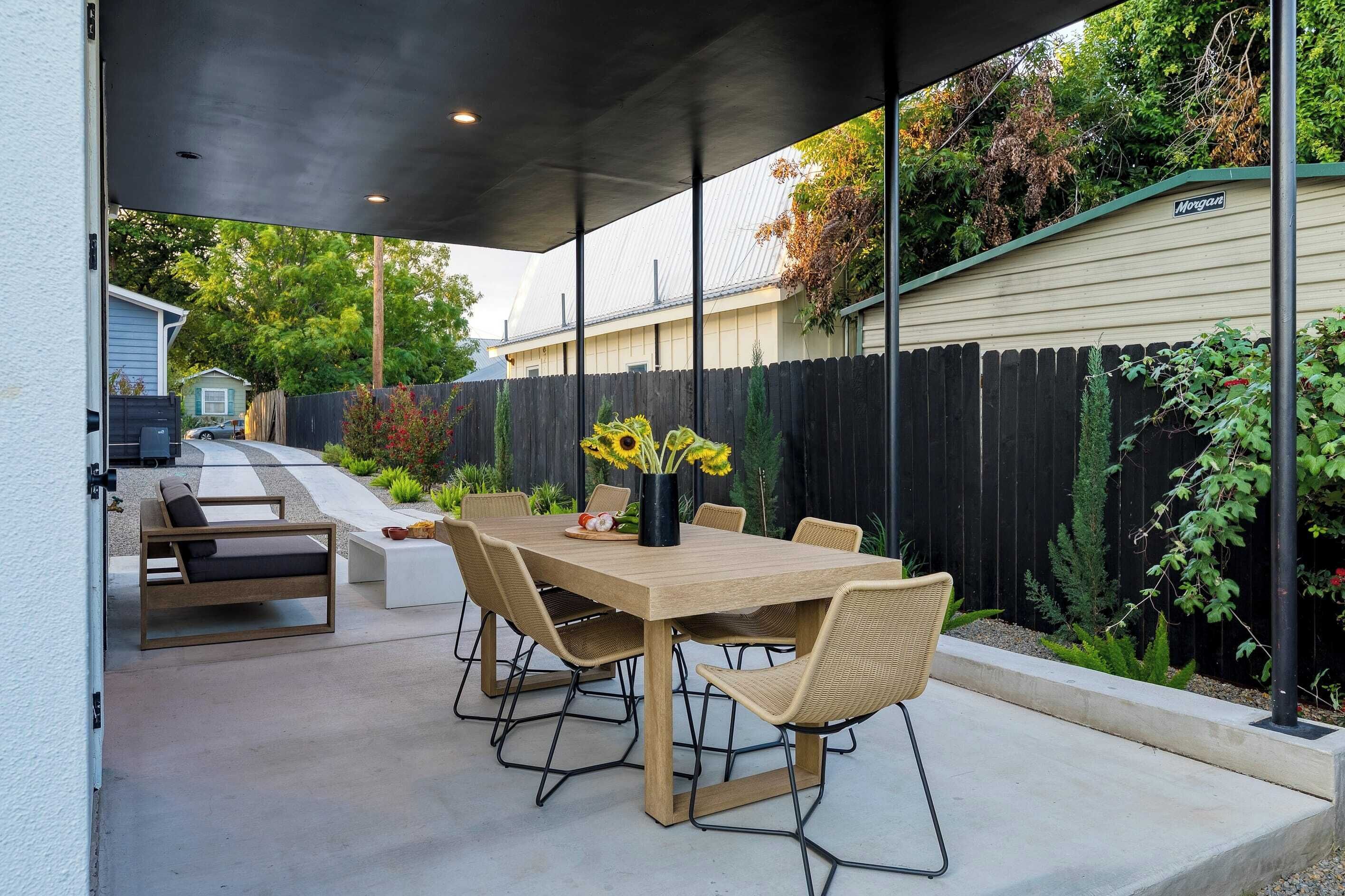 Een terras buiten met een houten tafel en stoelen, omgeven door een zwarte hek en begroeiing.