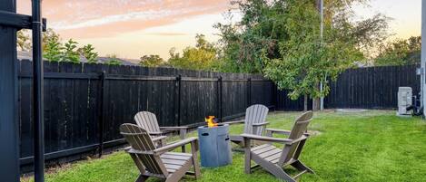 A backyard with a wooden fence, a fire pit, and Adirondack chairs.