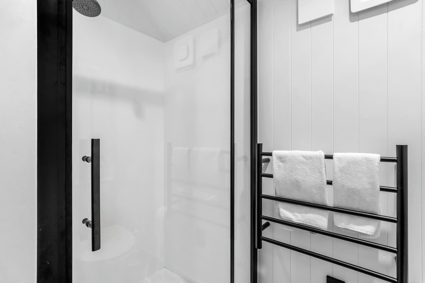 Une salle de bain moderne avec une douche à l’italienne en verre, un porte-serviettes noir avec trois serviettes et un mur blanc orné d’un petit miroir.