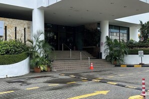 Entrada d'hotel amb disseny modern, amb escales cap a la llotja, una zona enjardinada amb plantes en testos i un camí ben marcat amb línies grogues.