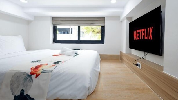 A hotel room with a bed, a window with curtains, a wall-mounted TV displaying the Netflix logo, and a wooden floor. - Siris Mia Residence - Self Check In (Ho Chi Minh City)