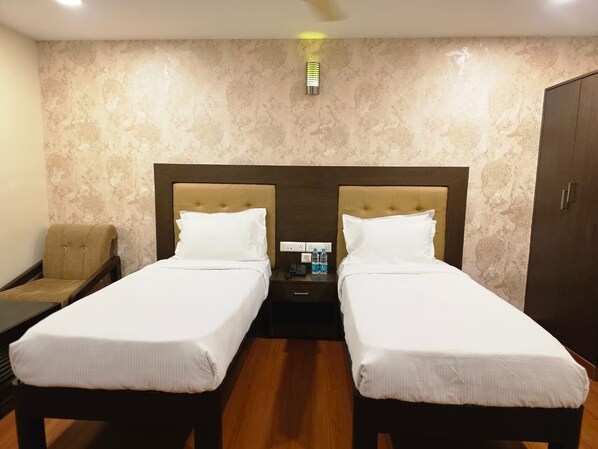 A hotel room with two single beds, a nightstand with a phone and water bottles, a chair, and a wardrobe. - MG Beatle Hotel Tirupati (Tirupati)