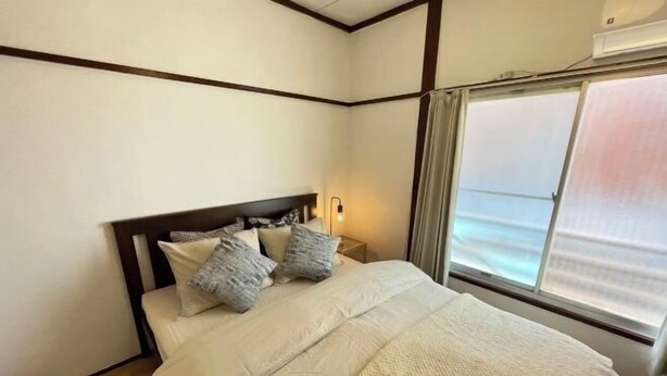 Una cama bien hecha con cabecera, una lámpara de noche y una ventana con cortinas.