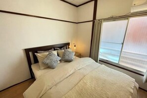 A neatly made bed with a headboard, a bedside lamp, and a window with curtains. - Wafu Stay · Kyomachiya (Tokyo)