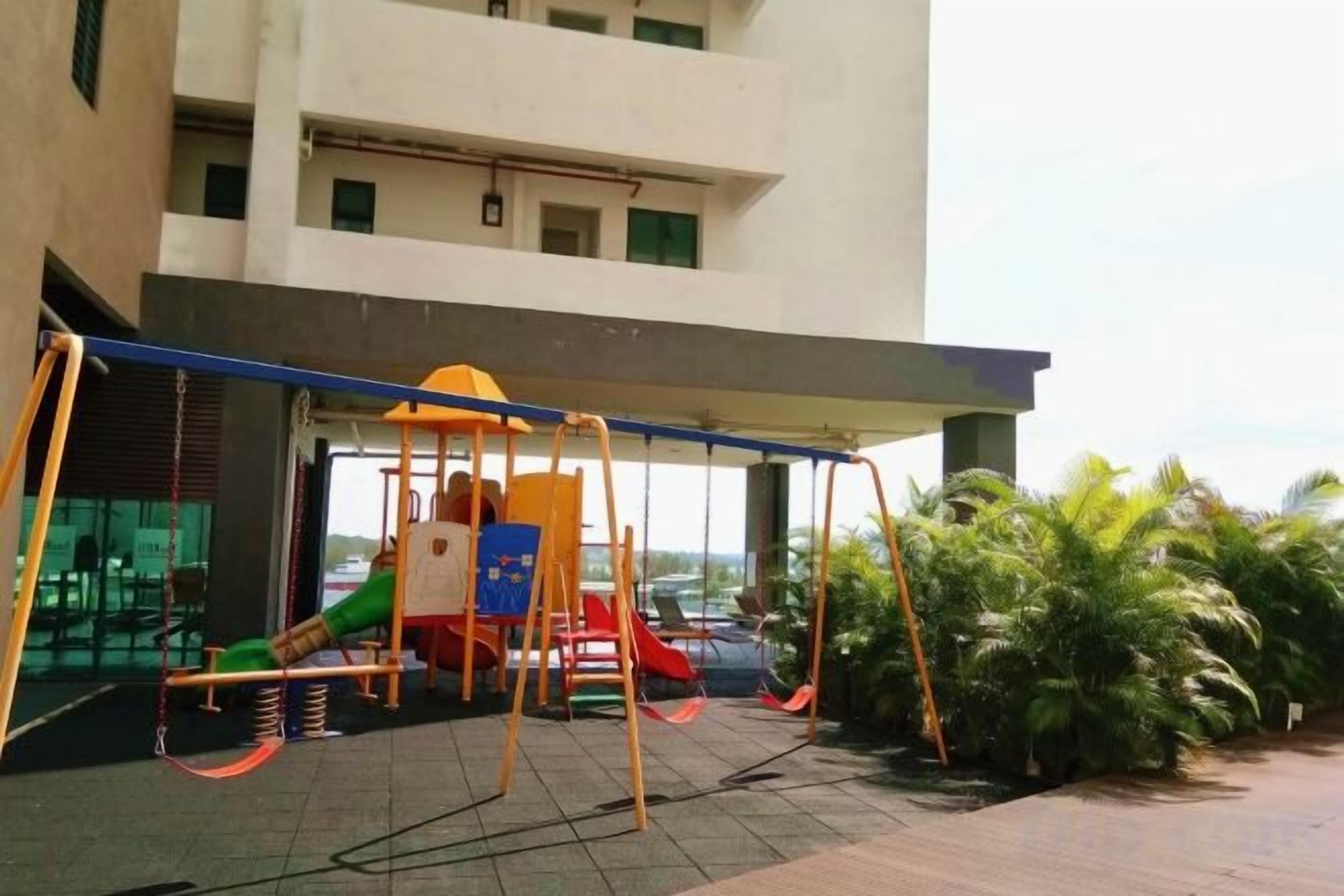 Zona de juegos al aire libre con columpios y toboganes, un área de descanso techada y vista a un edificio de varios pisos.