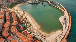 Vue aérienne d’une zone côtière comprenant une plage, un port de plaisance et des immeubles résidentiels aux toits recouverts de tuiles rouges.