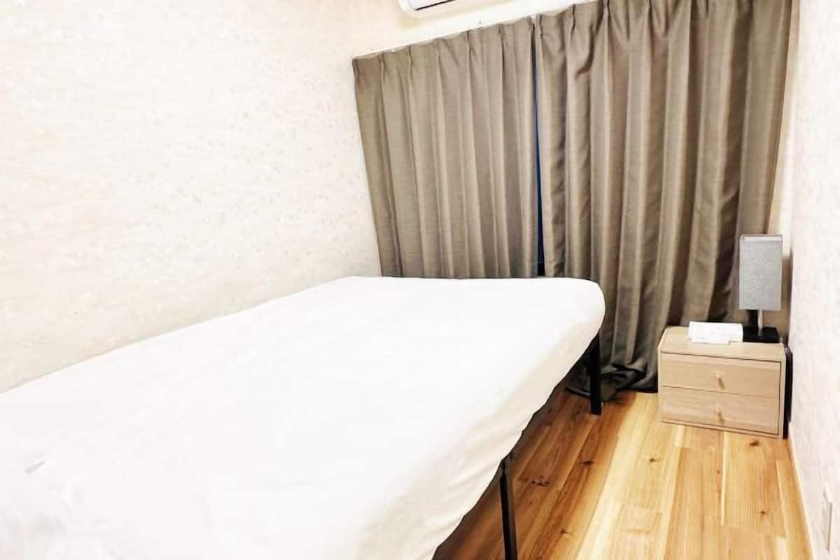 A small, single-bed room with a wooden floor, a bedside table with a lamp, and a curtain covering the window.
