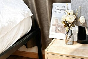 Un lit avec un drap blanc, une table de chevet ornée d’un vase de fleurs et un tableau encadré avec une inscription.