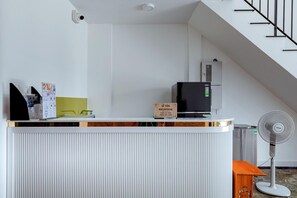Een receptiebalie met een bord waarop in- en uitchecktijden staan aangegeven, een zwart scherm, een Toshiba ventilator en een trap op de achtergrond.