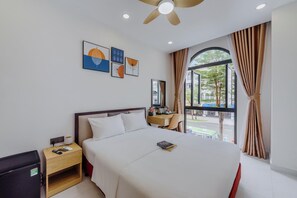 A hotel room with a bed, bedside table, a desk with a chair, a ceiling fan, and artwork on the wall. - Lalisa Hotel (Phu Quoc)