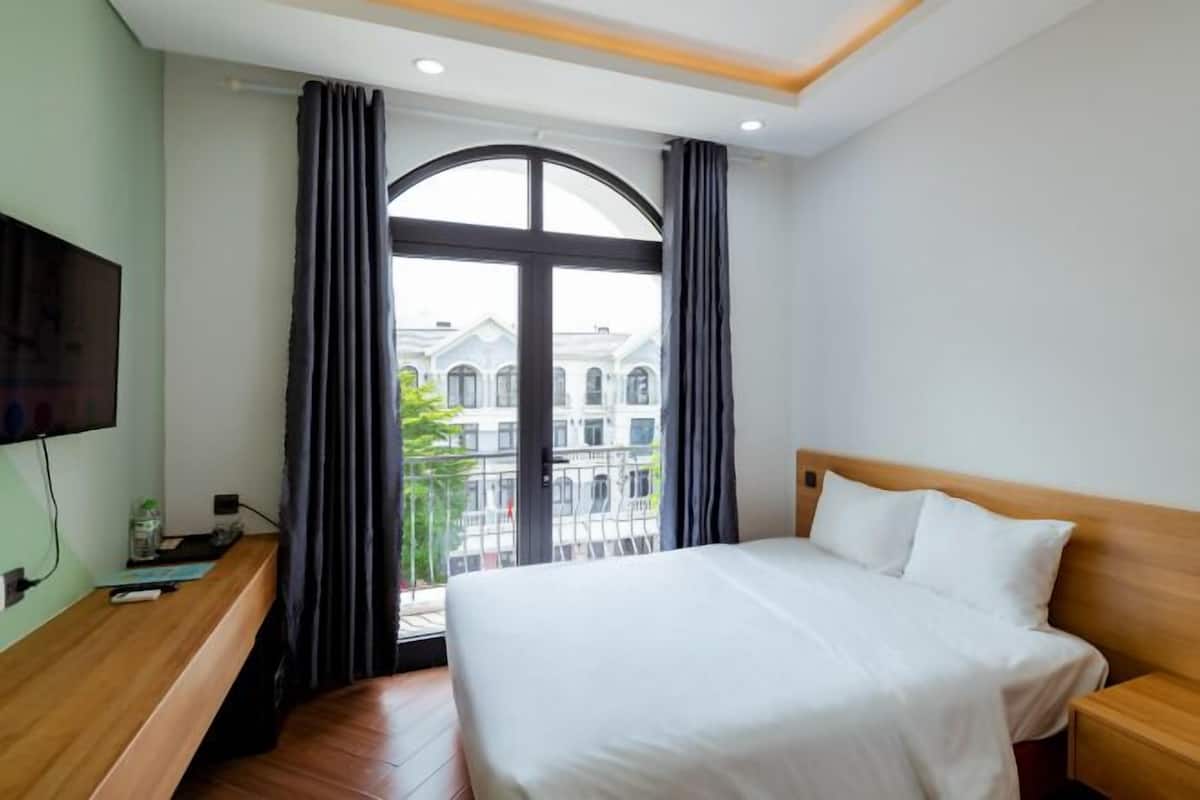 A hotel room with a bed, a TV, a wooden bench, a window with curtains, and a view of buildings.