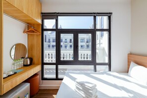 A hotel room with a bed, a wooden desk, a mirror, and a large window with a view of buildings. - Lalisa Hotel (Phu Quoc)