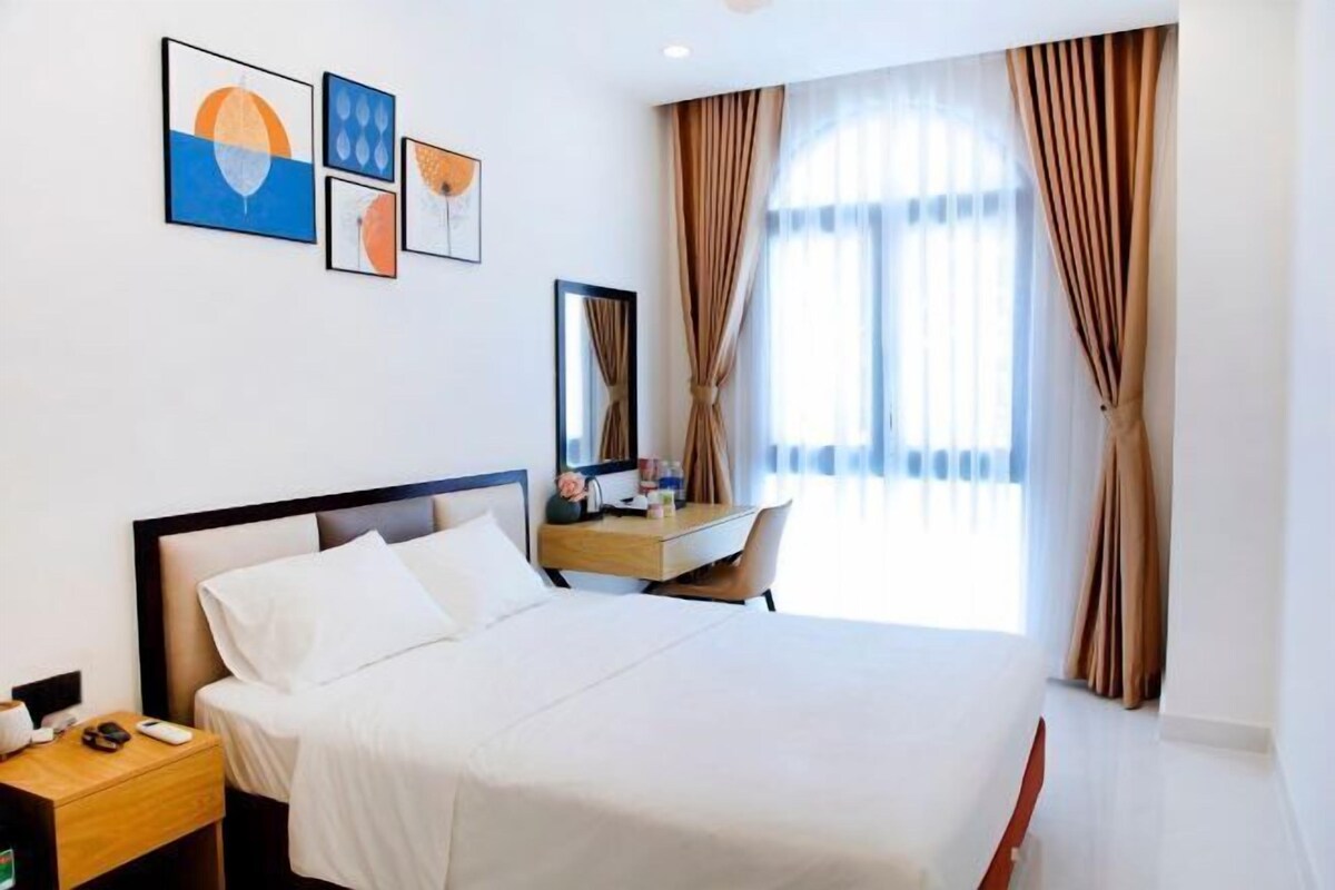 A hotel room with a bed, bedside table, desk, chair, and large window with curtains.