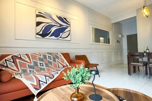 A modern living room with a sofa, a coffee table with a vase, a painting on the wall, and a dining area in the background. - Nilai Vision City 4+1 PAX Akasia Suite (Nilai)