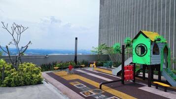 A rooftop playground with a slide and climbing structures, surrounded by greenery and a view of the city.
