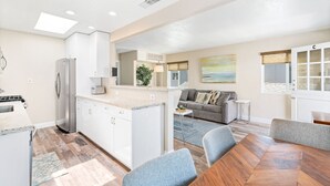A modern kitchen with white cabinets, a built-in oven, and a refrigerator. There is a dining area with a wooden table and blue chairs. A gray sofa and a small side table are also visible. - 5BR Modern Retreat, One House from the Sand with AC, Patios & Upper Deck! (Newport Beach)