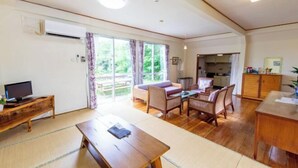 Un vaste salon comprenant une table en bois, un canapé et des fauteuils, un téléviseur, et un coin cuisine à l’arrière-plan.