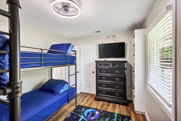 A bunk bed room with a ceiling fan, a TV, a dresser, and a window with blinds.