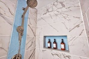 Un baño con ducha, pared de azulejos azules y estante con tres botellas.