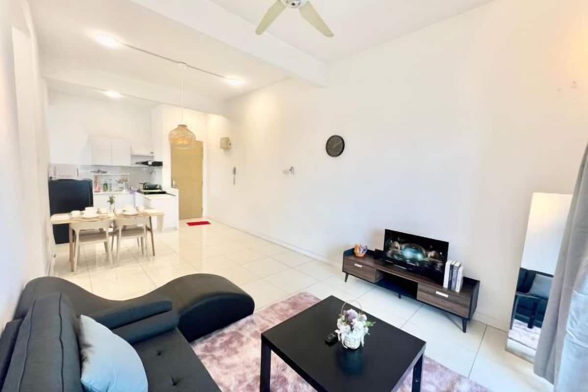Un salon moderne avec un canapé, une table basse et un téléviseur. Au fond, on aperçoit une cuisine avec un coin repas.