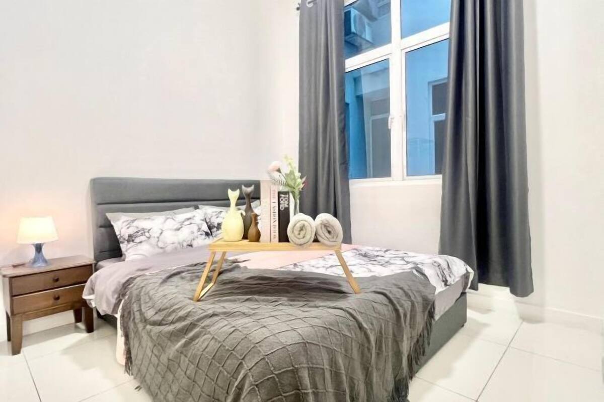 A neatly made bed with a gray quilt, a bedside table with a lamp, and a window with curtains.