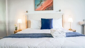 Una cama bien hecha con ropa blanca y azul, dos lámparas de noche y un cuadro sobre la cama.