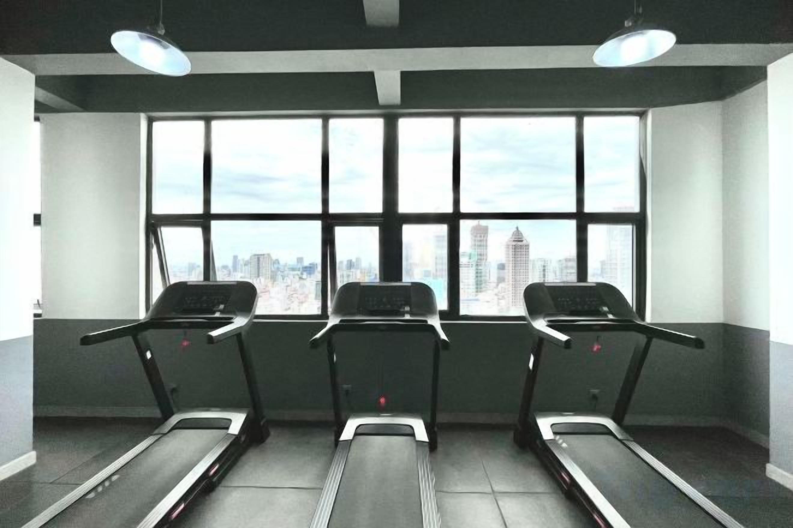 A gym with treadmills aligned against a wall with large windows, overlooking a cityscape.