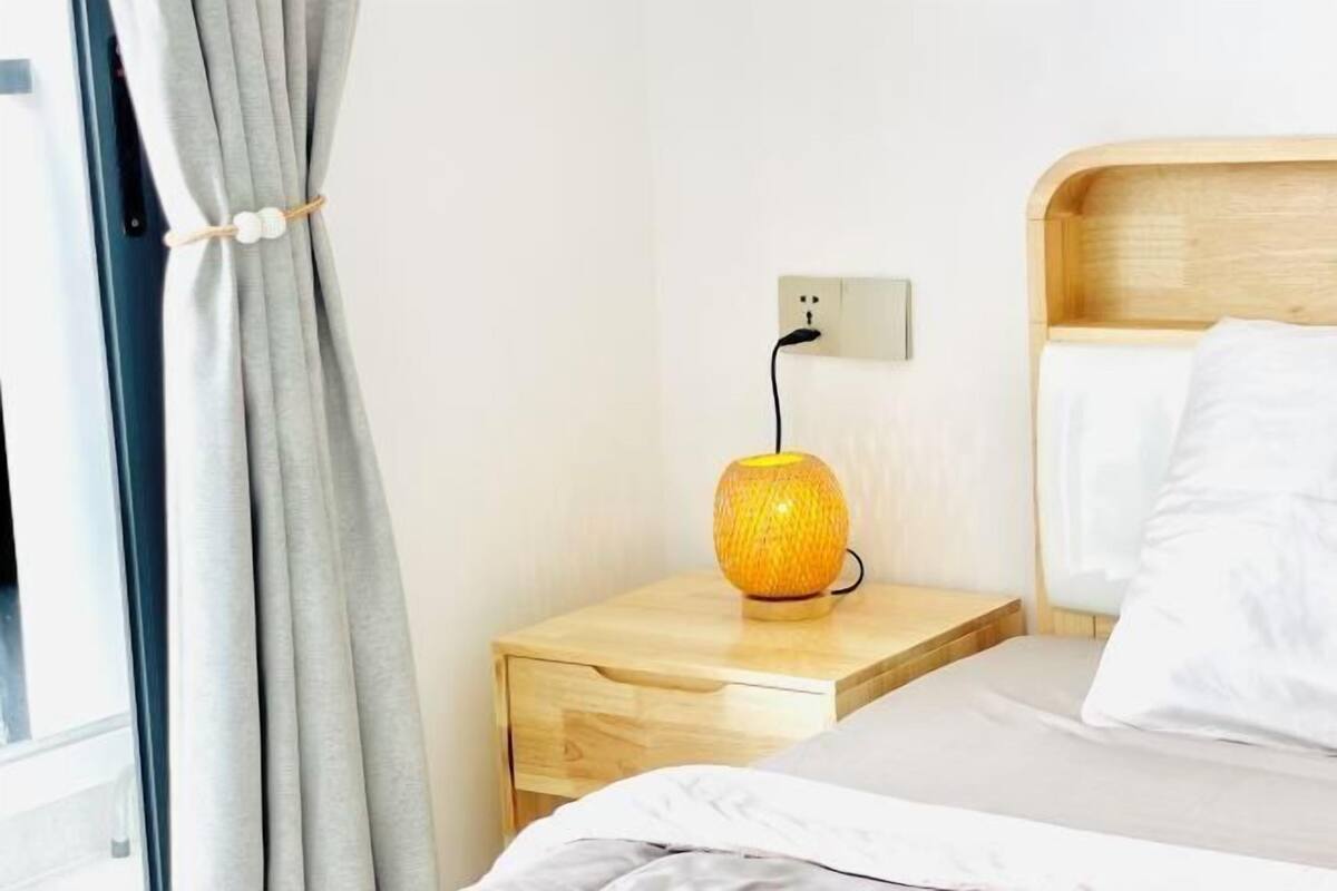 Una cama bien hecha con ropa blanca, una mesita de noche de madera con una lámpara y un enchufe en la pared.