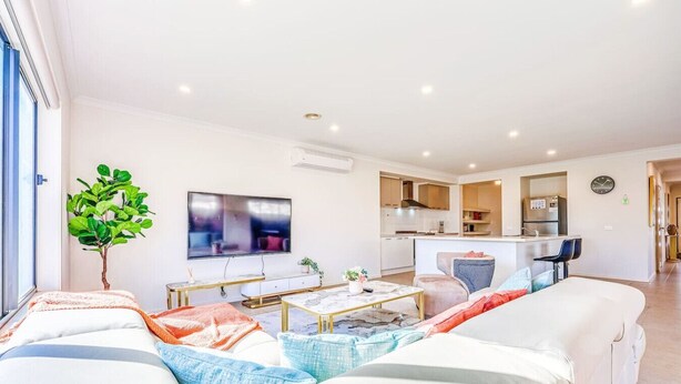 A modern living room with a sofa, coffee table, and a TV. There is a kitchen area with cabinets and a dining table. - StayAU 4BR Tranquil Family Escape House Point Cook (Point Cook)