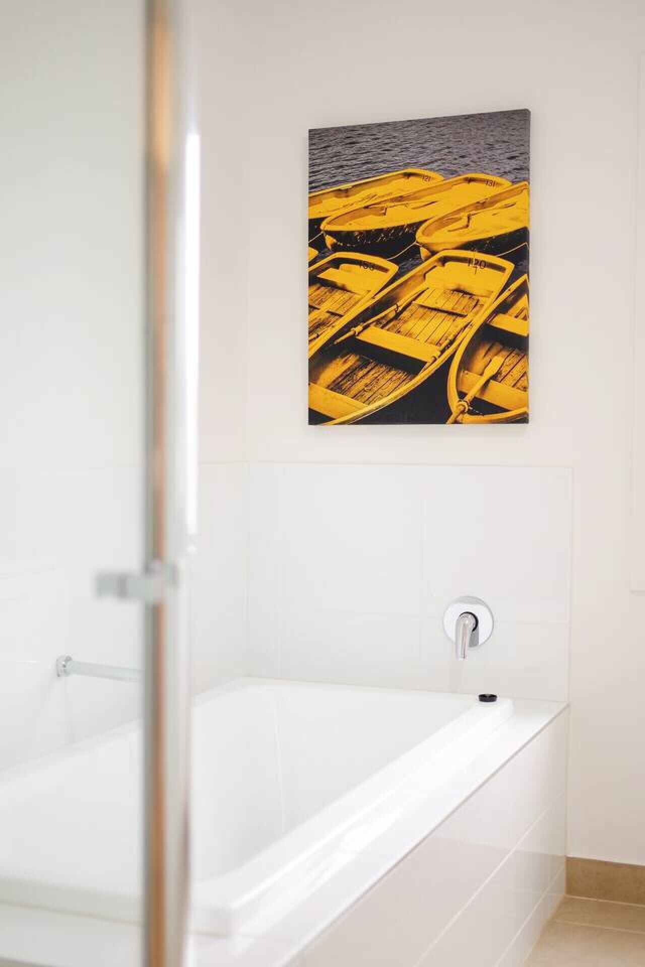 Une salle de bain moderne avec une baignoire blanche, une douche en verre et une œuvre d’art jaune représentant des bateaux sur une plage.