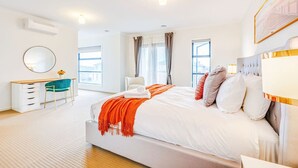 A modern bedroom with a large bed, a desk, and a chair. There are windows with curtains, a round mirror, and a framed picture on the wall. - StayAU 4BR Tranquil Family Escape House Point Cook (Point Cook)