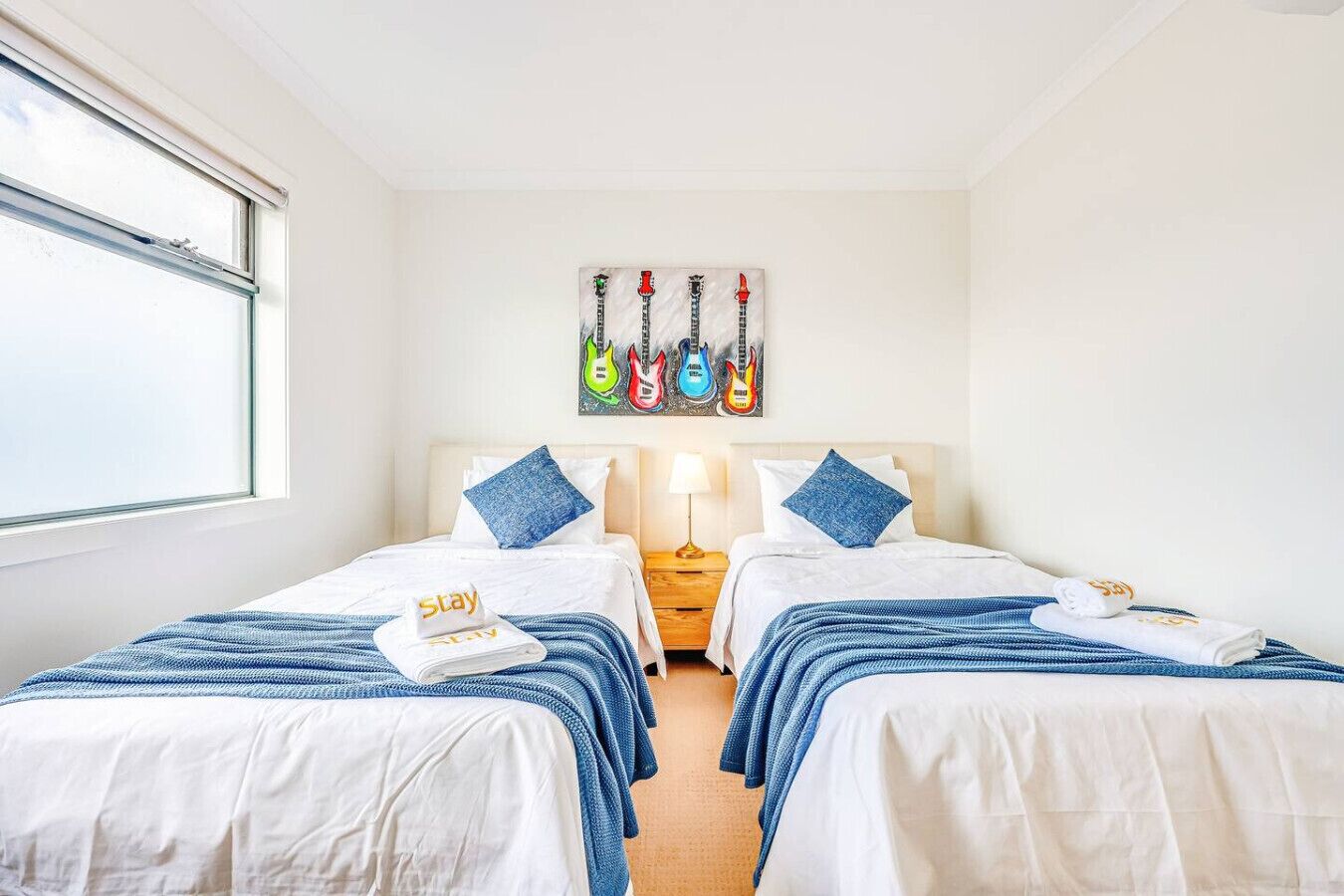 Habitación de hotel con dos camas, una ventana, un cuadro de guitarras y una mesita de noche con una lámpara.