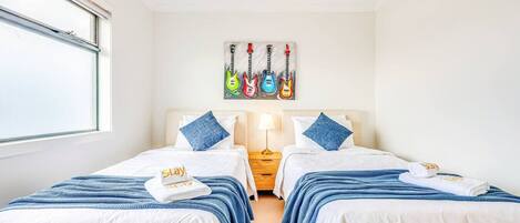 Habitación de hotel con dos camas, una ventana, un cuadro de guitarras y una mesita de noche con una lámpara.