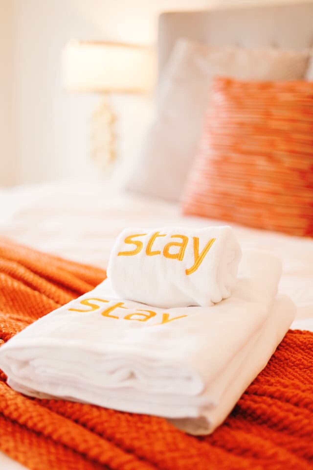Cama bem arrumada com uma toalha branca dobrada e um travesseiro branco com a palavra 'stay' bordada em amarelo.