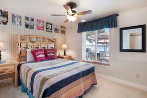Une chambre à coucher avec une tête de lit en bois, une couvre-lit matelassée, deux lampes de chevet, un ventilateur de plafond et une fenêtre avec des stores.