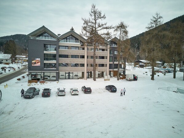 A multi-story building with a modern architectural design, featuring large windows and a parking area with several cars.
