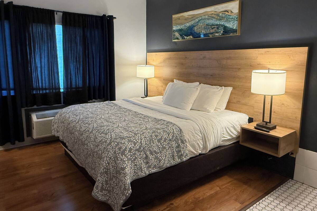 Une chambre d’hôtel avec un lit, deux lampes de chevet, une tête de lit en bois, un tableau au mur et une fenêtre avec des rideaux.