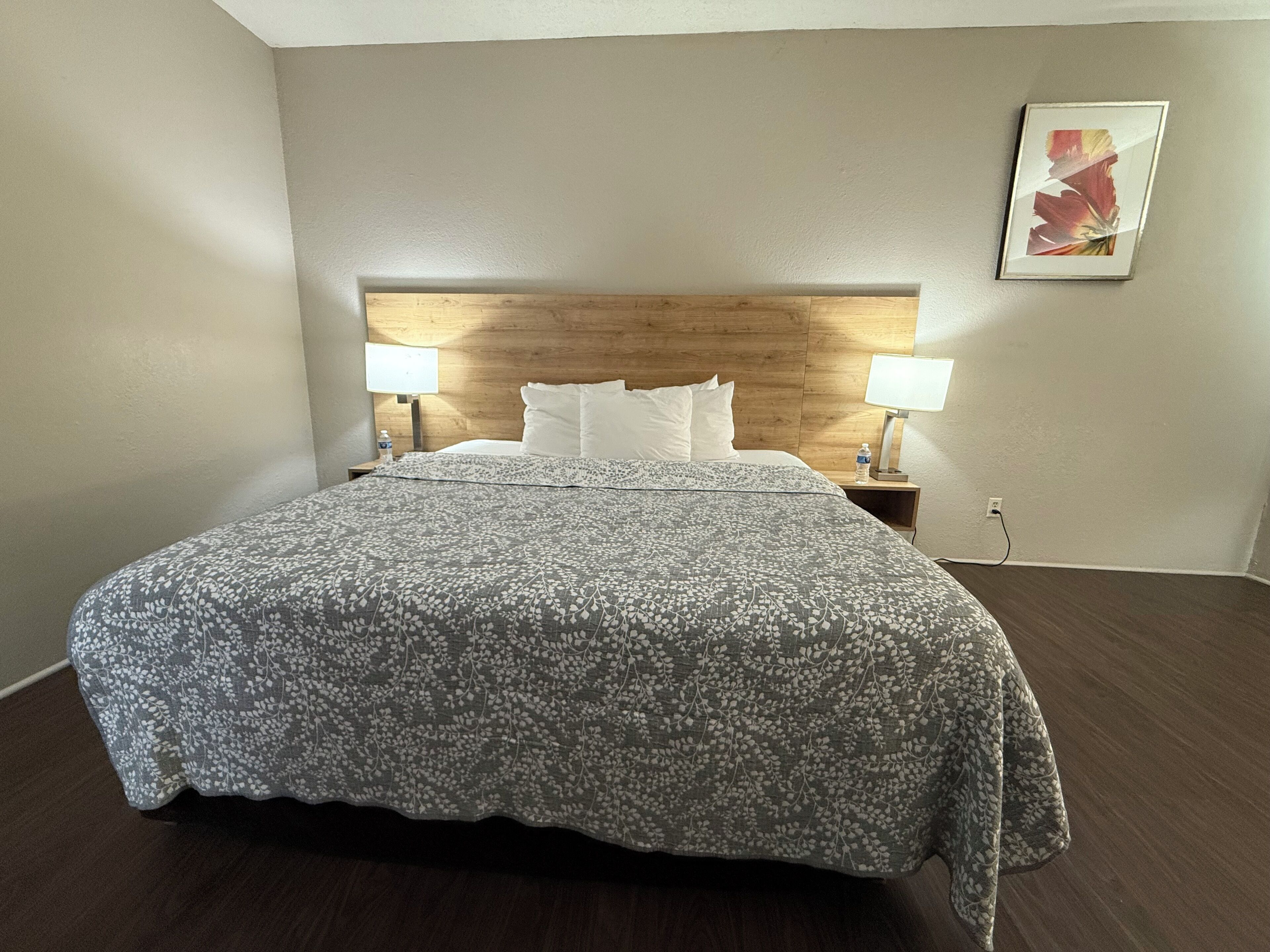 A hotel room with a wooden headboard, a floral-patterned bedspread, two bedside lamps, and a framed artwork on the wall.