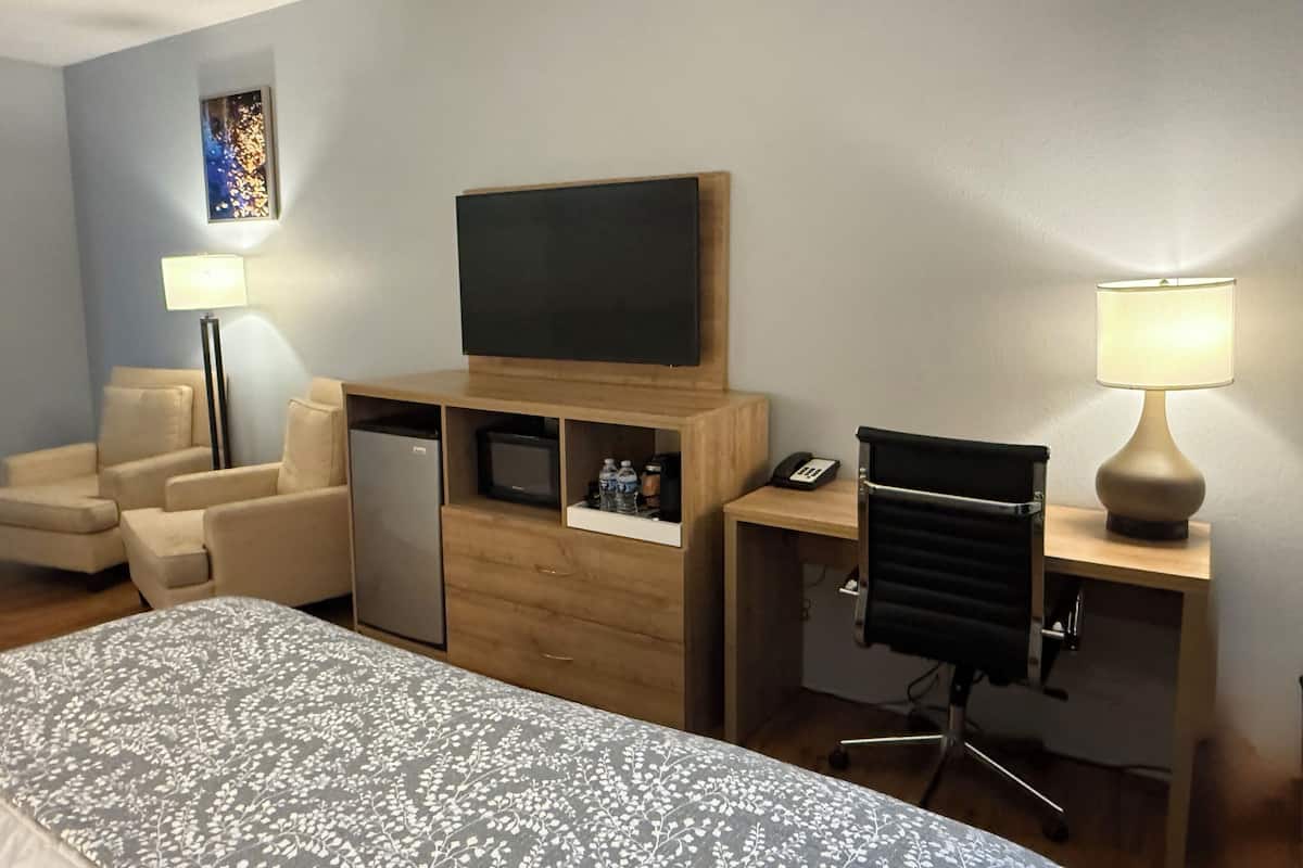 Une chambre d’hôtel comprenant un lit, un bureau avec une chaise, un téléviseur à écran plat, un réfrigérateur, une lampe et une petite étagère avec quelques objets.