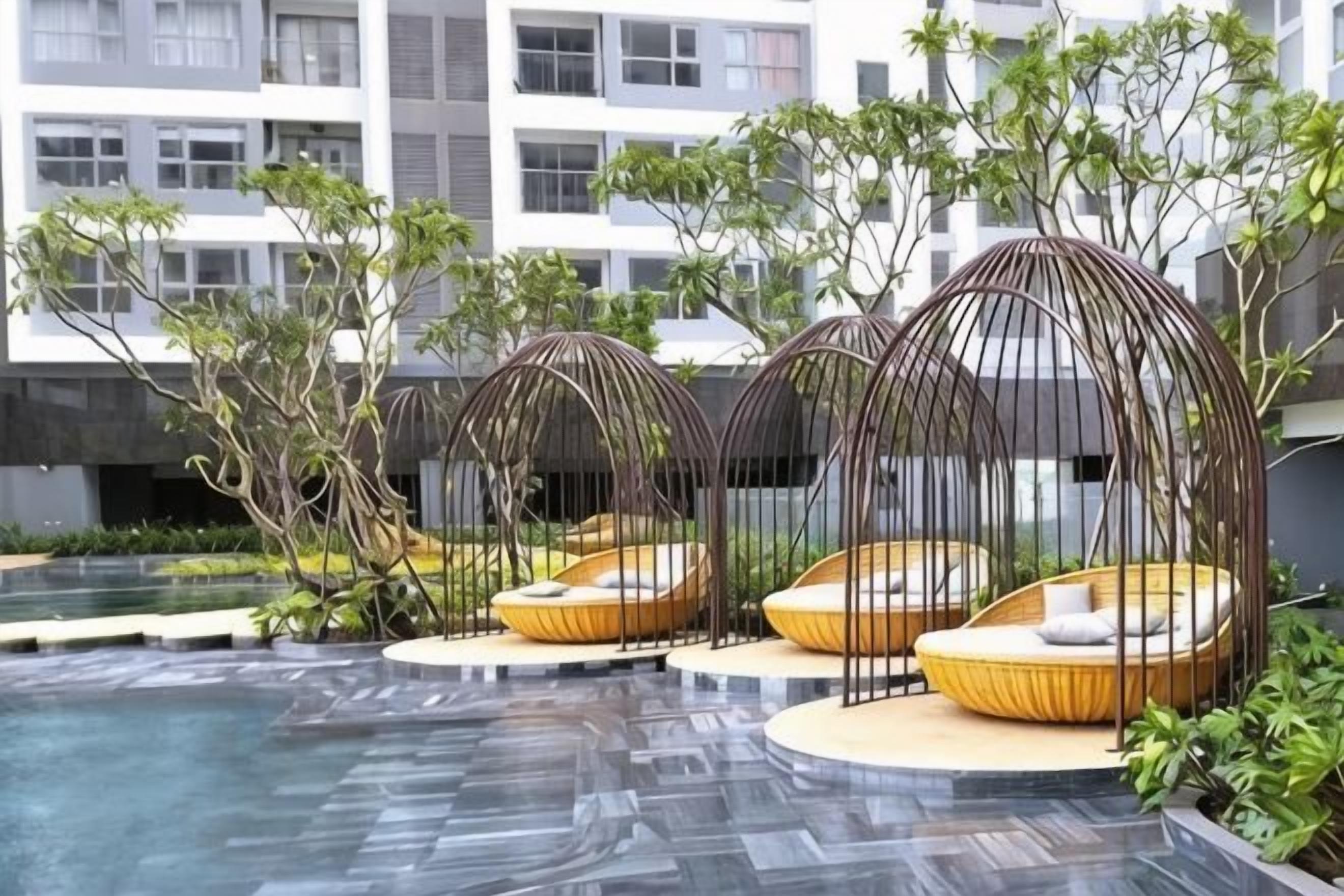 Zona de piscina con tres sillones de mimbre bajo una pérgola, rodeada de vegetación y un edificio moderno al fondo.