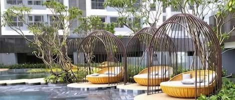 Zona de piscina con tres sillones de mimbre bajo una pérgola, rodeada de vegetación y un edificio moderno al fondo.