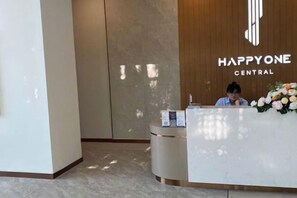 A reception desk with a logo, a woman behind it, and a wall with a vertical design. - Happy One Central - Apartment Thu Dau Mot (Thu Dau Mot)